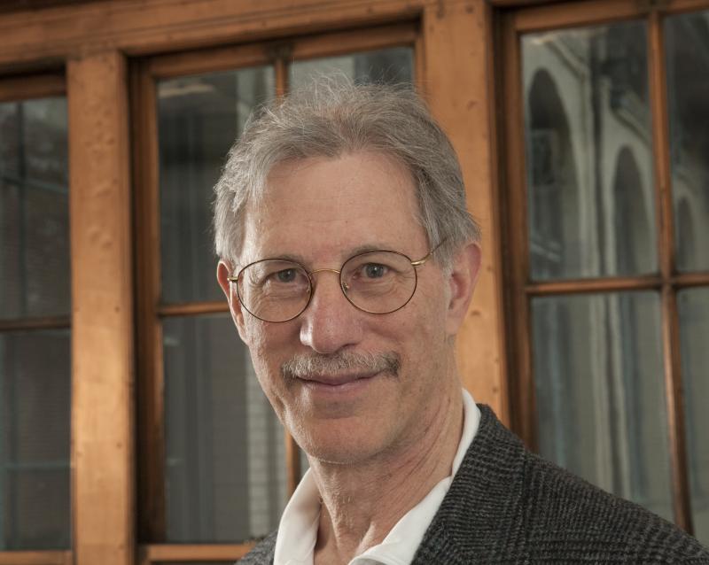An older man with glasses and gray hair smiles in front of wooden-framed windows.