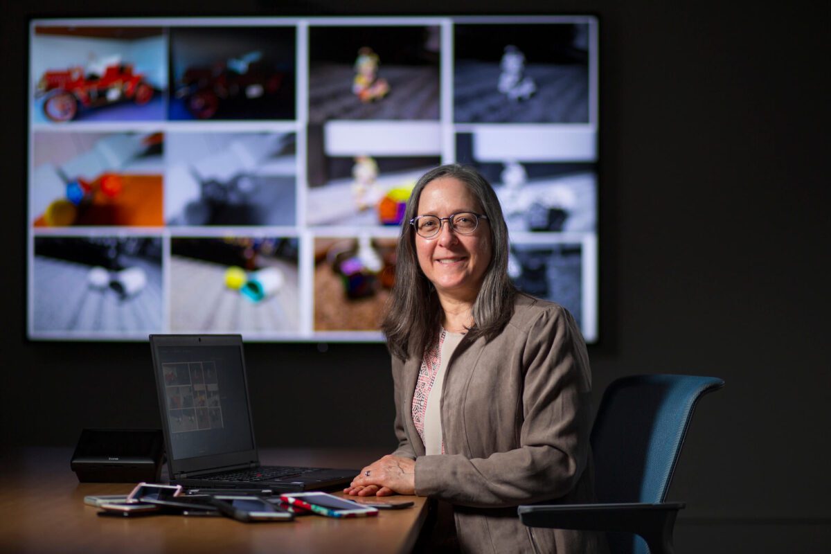 Jennifer Newman at a desk with a laptop; blurry photos displayed on a screen behind her.