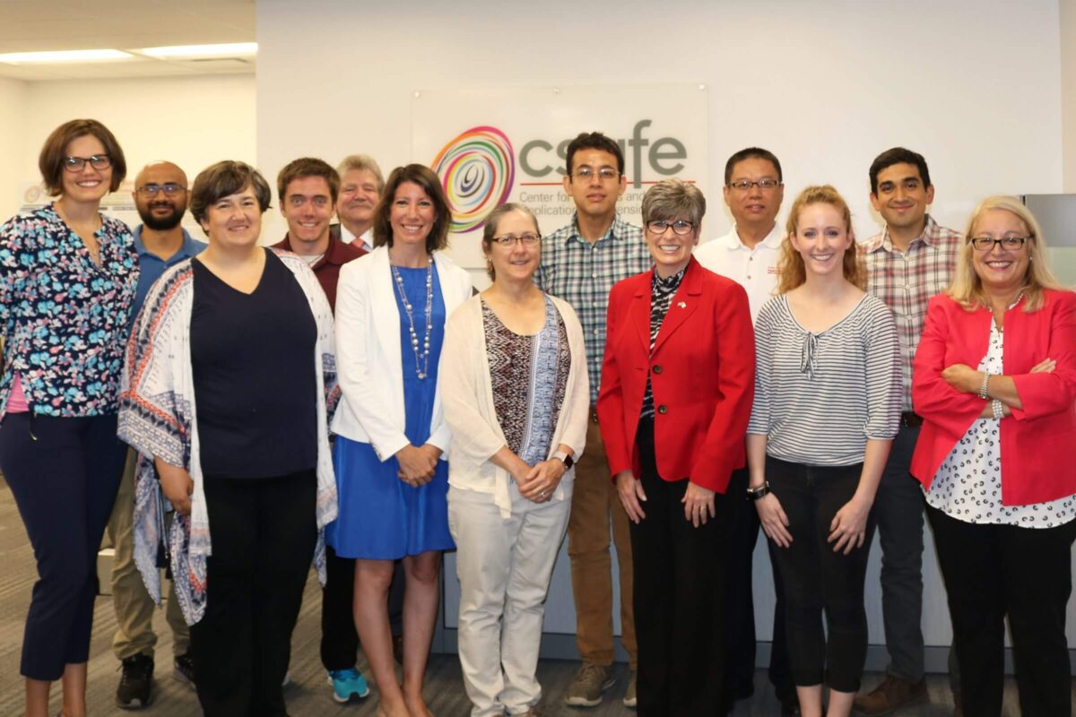 A group of thirteen people smiling and posing together in an office setting near a sign that says "ccafe.