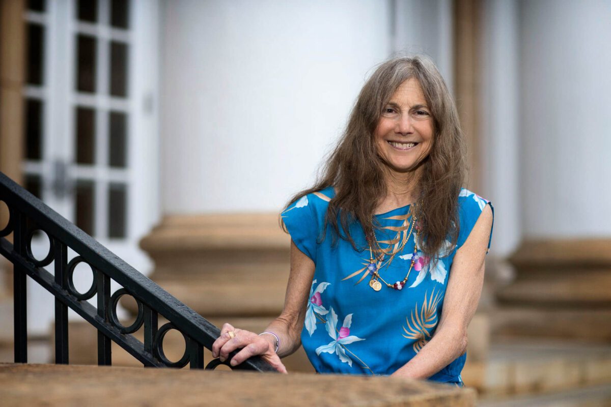A smiling woman in a blue floral top stands outside by a railing, with columns in the background.