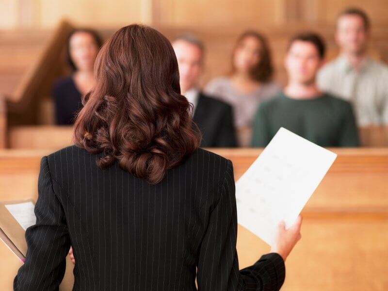 A woman in a suit holding papers stands in a courtroom facing a seated jury.