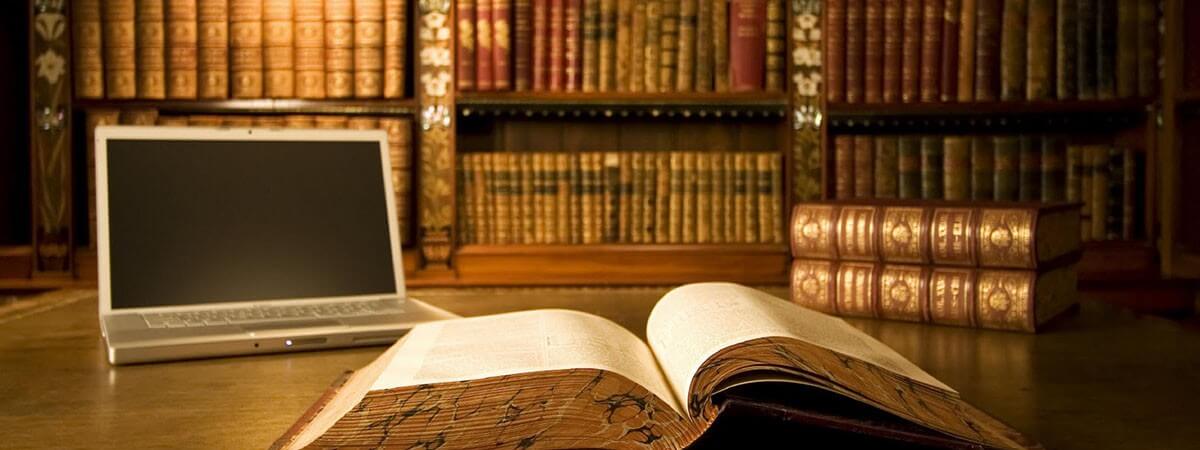 Open book on a desk with a laptop and stacked books, in front of shelves filled with old books.