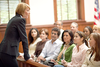 A lawyer speaks to a seated jury in a courtroom, with people listening attentively.