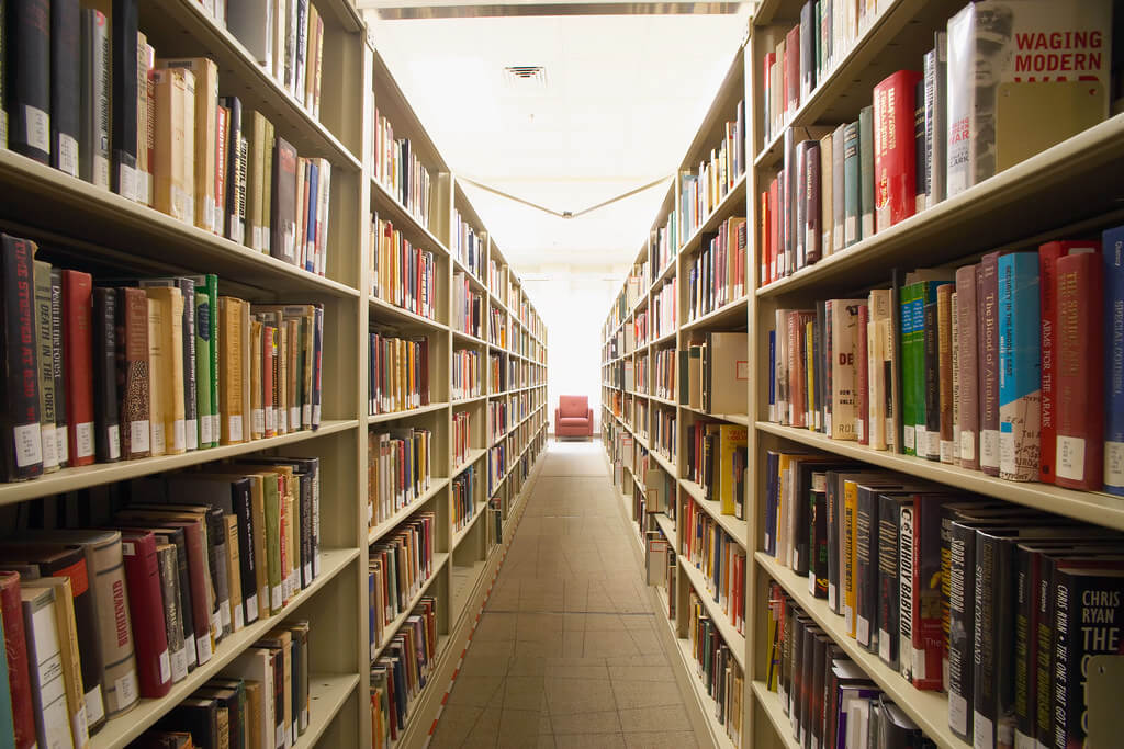 Rows of bookshelves in a library with a single chair at the brightly lit end of the aisle.