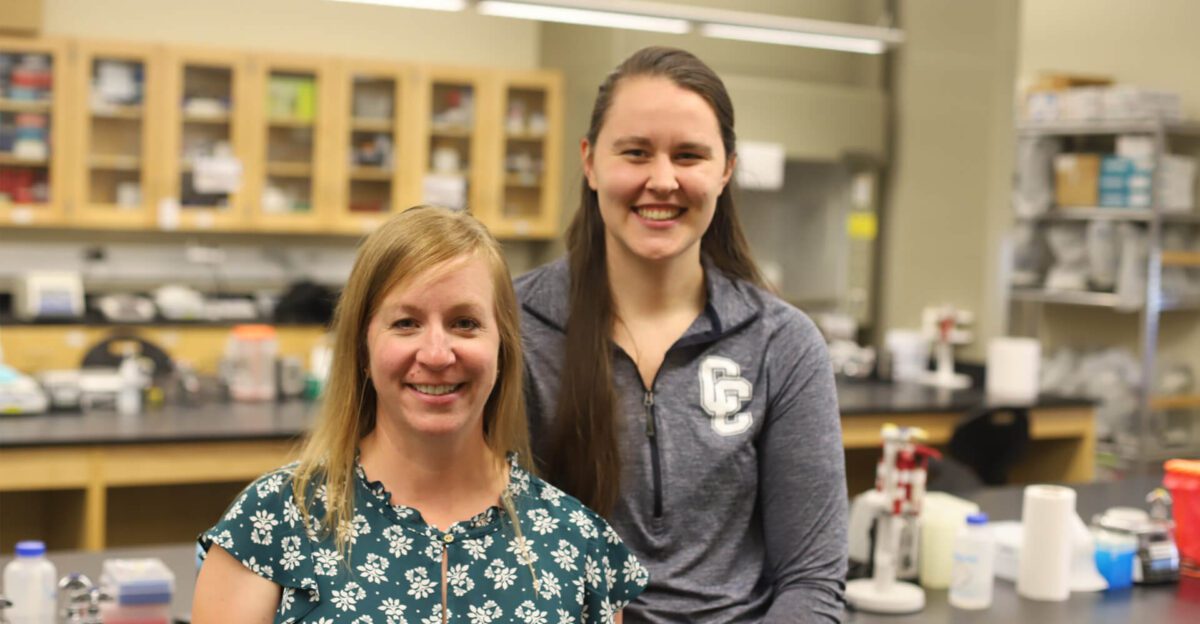 Two women smiling in a science lab with shelves and equipment in the background.