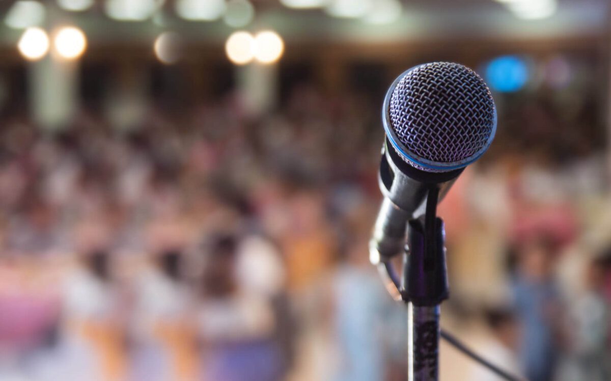 A microphone on a stand with a blurred crowd in the background, suggesting a public speaking event.
