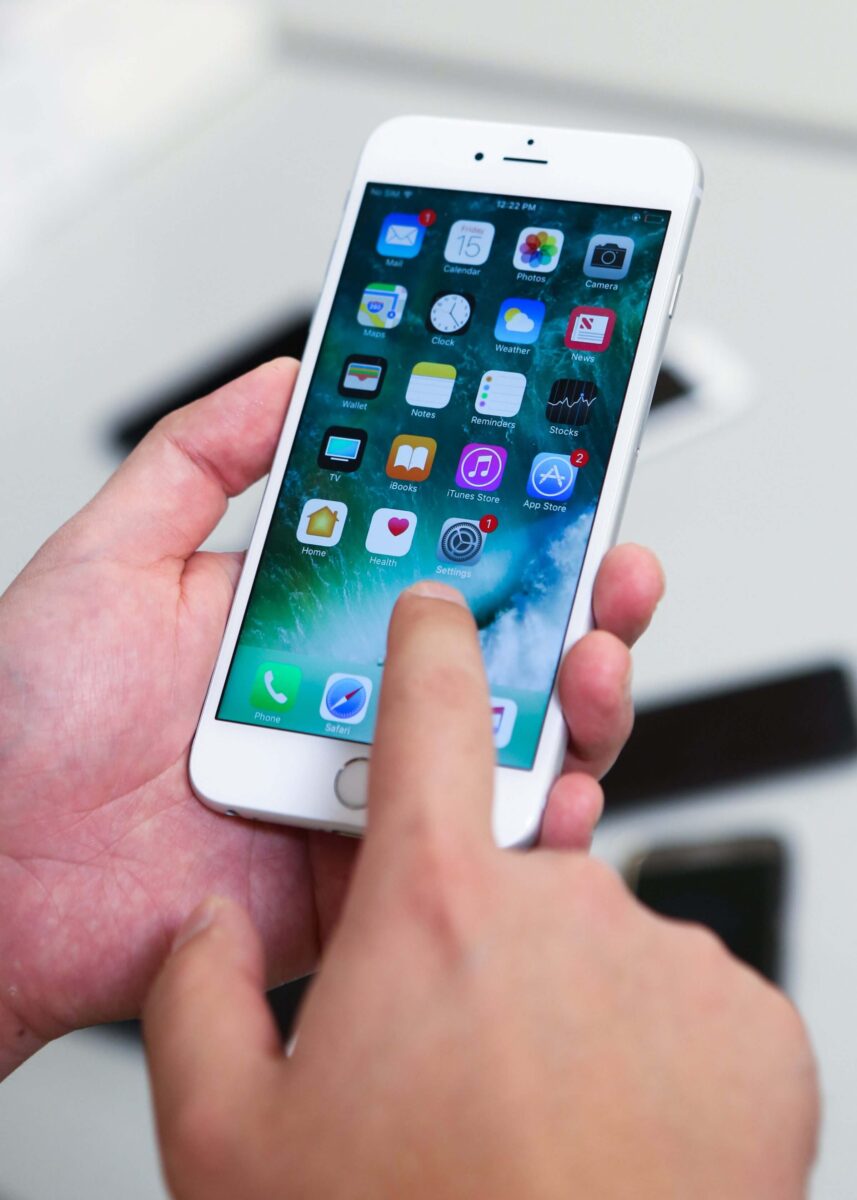 A person uses their finger to tap the screen of a white iPhone displaying app icons.