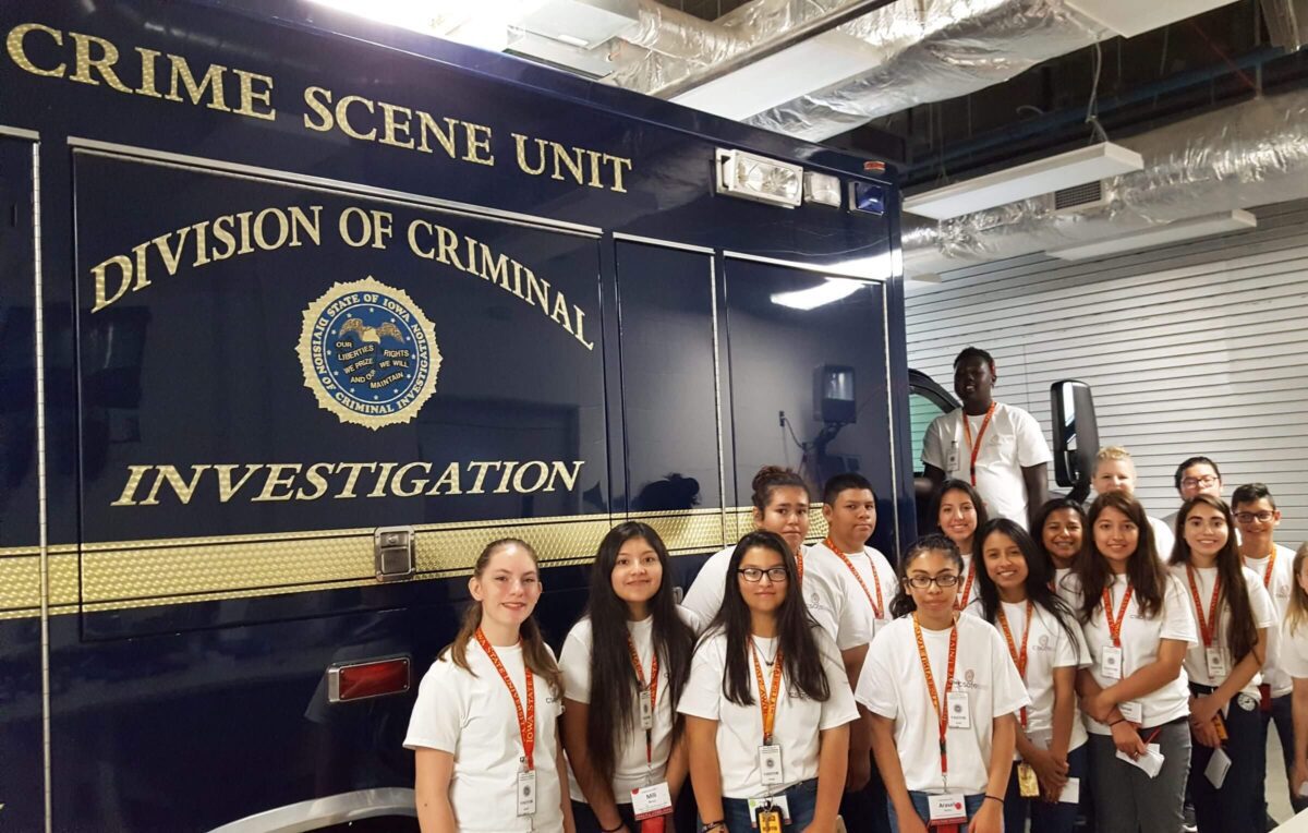 A group of students in white shirts stand by a Crime Scene Unit truck inside a building.
