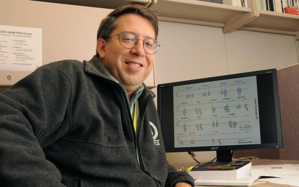 A man in glasses smiles at a desk with a computer showing a colorful data graph in an office.