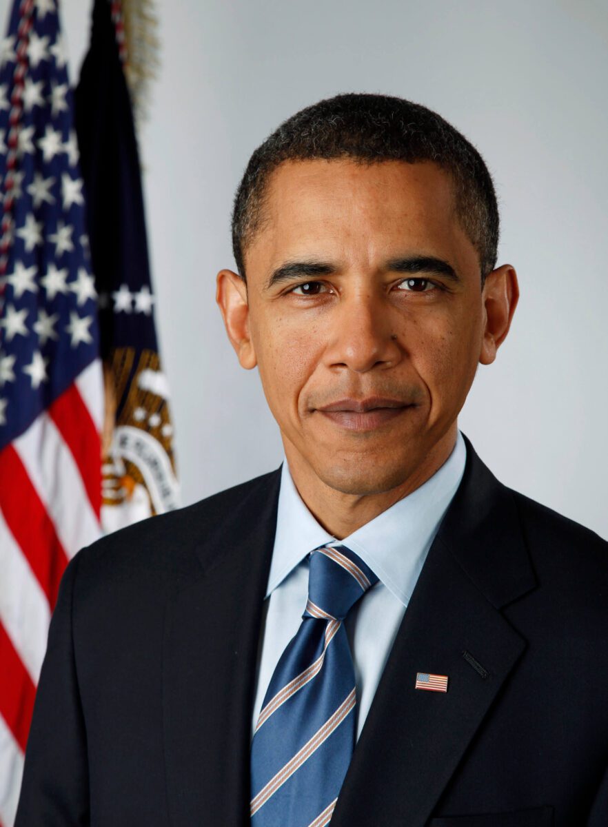 A man in a suit and tie stands in front of an American flag and official emblem.