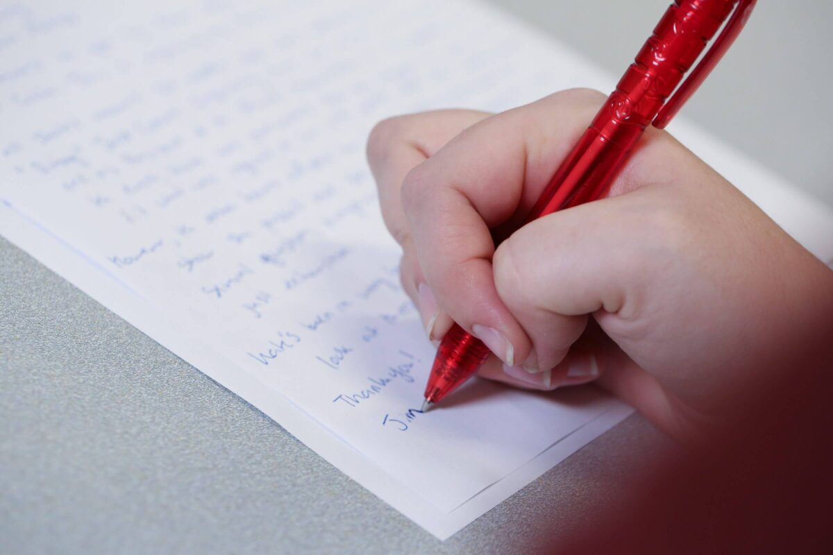 A person writing on lined paper with a red pen; handwriting is visible but mostly out of focus.
