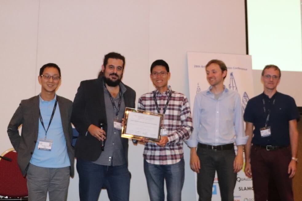 Five men standing together, one holding a framed certificate, all smiling at an indoor event.