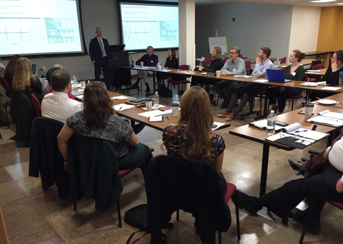 A group of people in a meeting room listen to a presenter standing by a screen with charts and data.