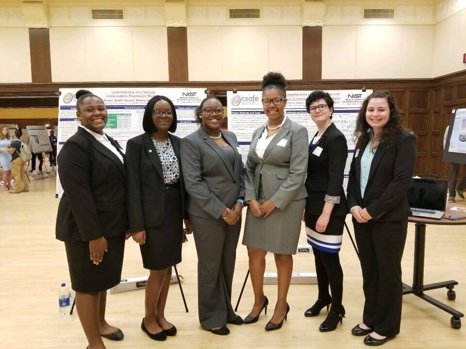 6 women in business attire stand and smile in front of research posters at a conference event.