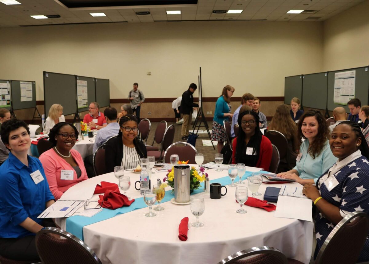 Six people sit smiling at a round table during a conference or seminar in a banquet hall.
