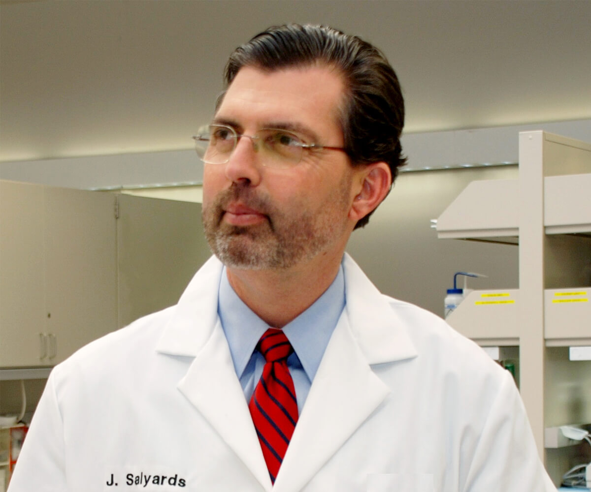 Man in a white lab coat and red striped tie standing in a laboratory, looking to the side.