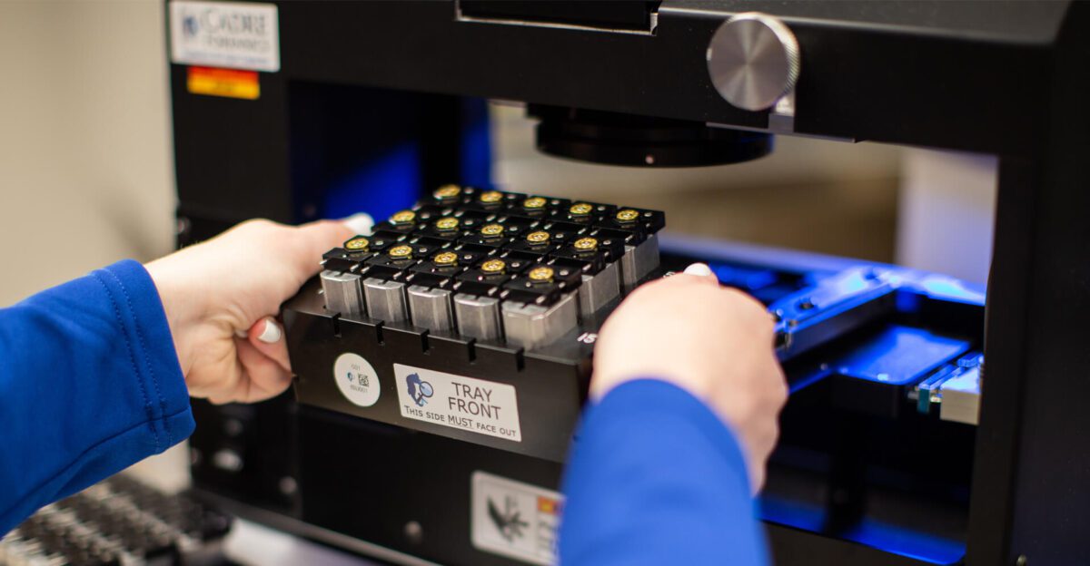 A person inserts a tray of test tubes into a laboratory machine for analysis.