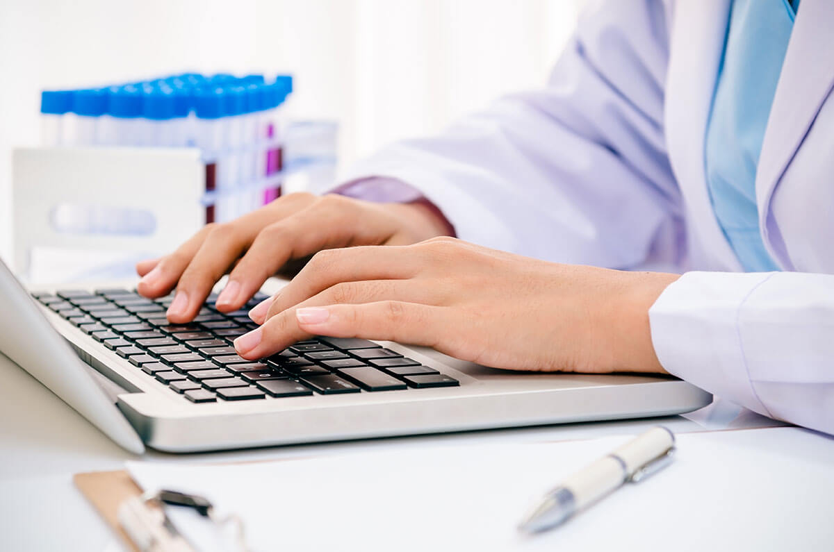 A person in a lab coat types on a laptop beside test tubes and a pen on a desk.