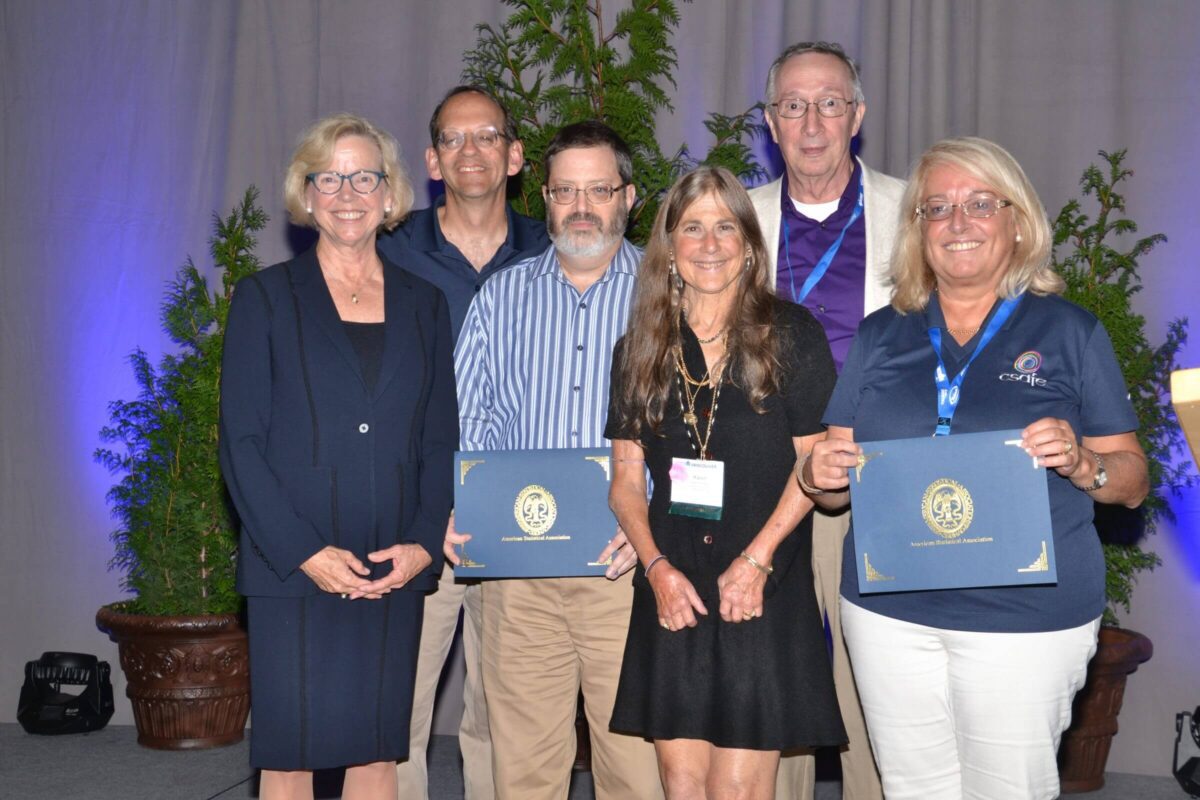 Six adults stand smiling; two hold certificates in front of a backdrop with green plants.