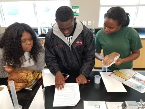 Three students work together in a science lab, reading papers and examining an object.