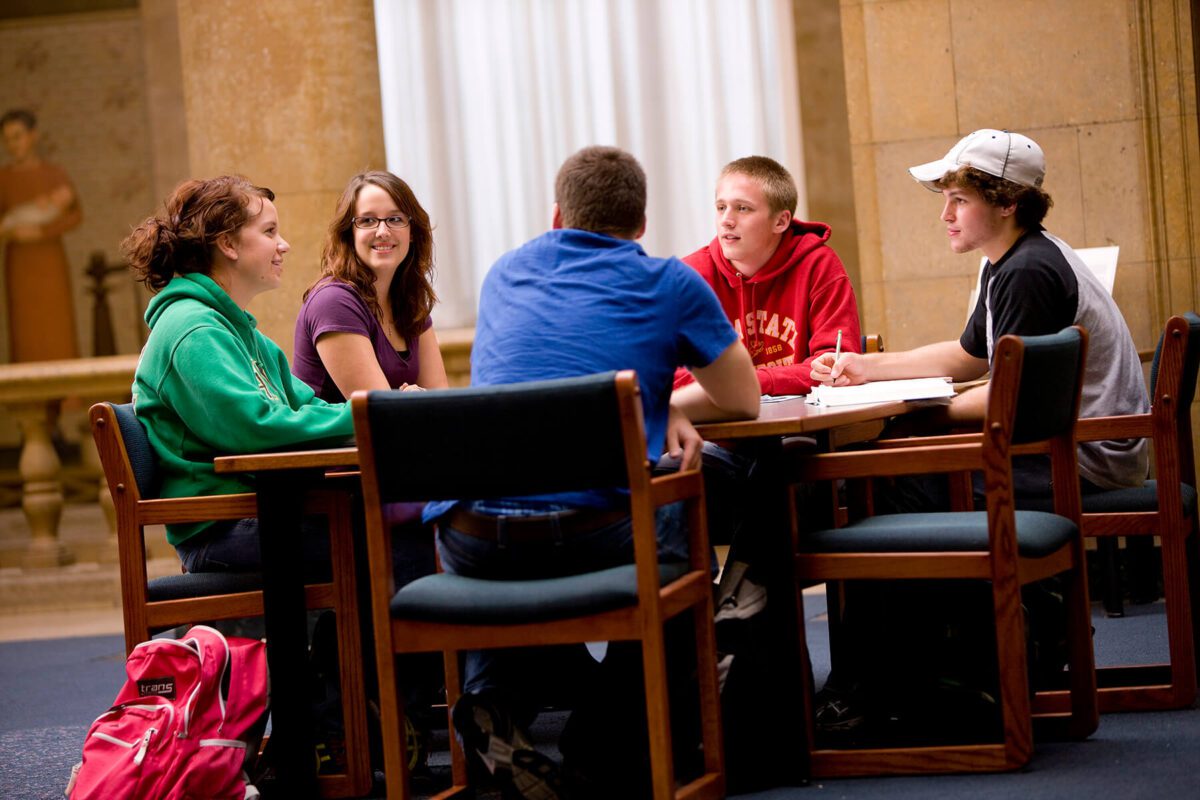 Five students sit around a table, studying and talking in a library or study area.