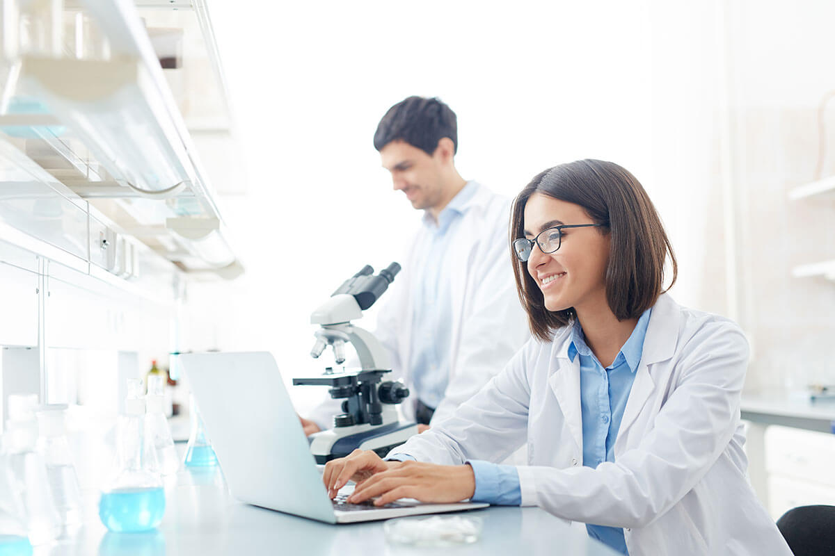 Smiling scientist using a laptop in a laboratory, with a colleague and microscope in the background.