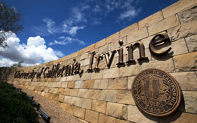 Stone wall with "University of California, Irvine" and the university seal under a partly cloudy sky.
