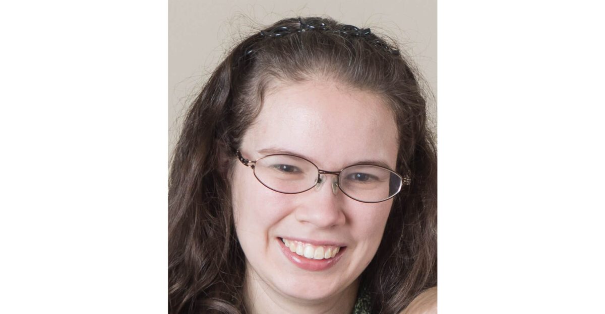 Woman with long brown hair, wearing glasses and smiling, in front of a plain light background.