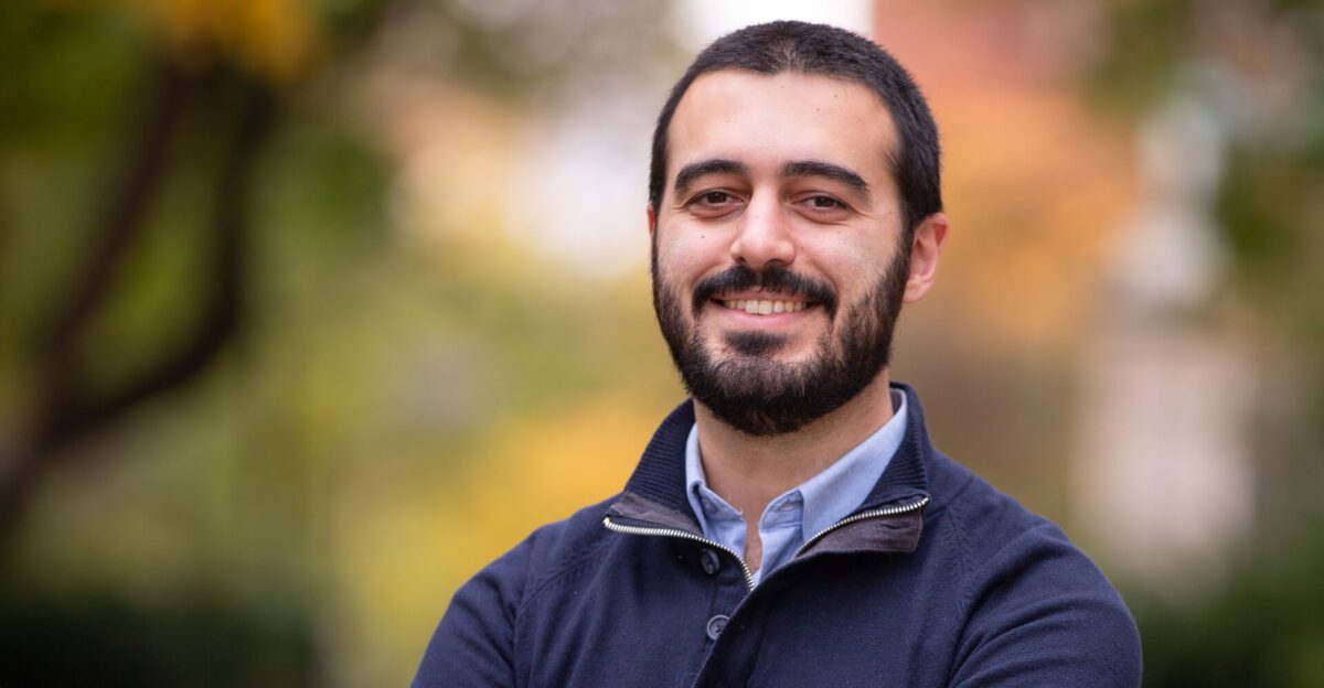 A man with a beard and dark hair smiles outdoors, wearing a navy sweater over a collared shirt.