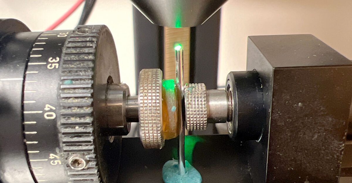 Close-up of scientific equipment with a green laser beam shining on a thin rod held by metal clamps.