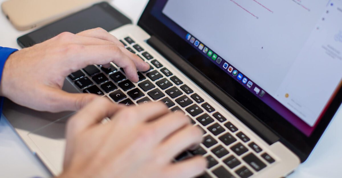 Hands typing on a laptop keyboard with a smartphone placed beside the laptop on the desk.