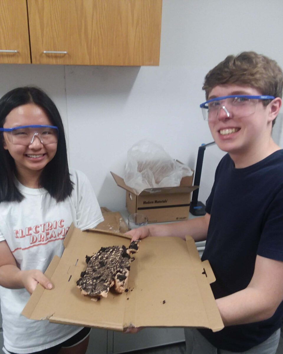 Two smiling teens in goggles hold a cardboard tray with a chocolate-covered dessert.